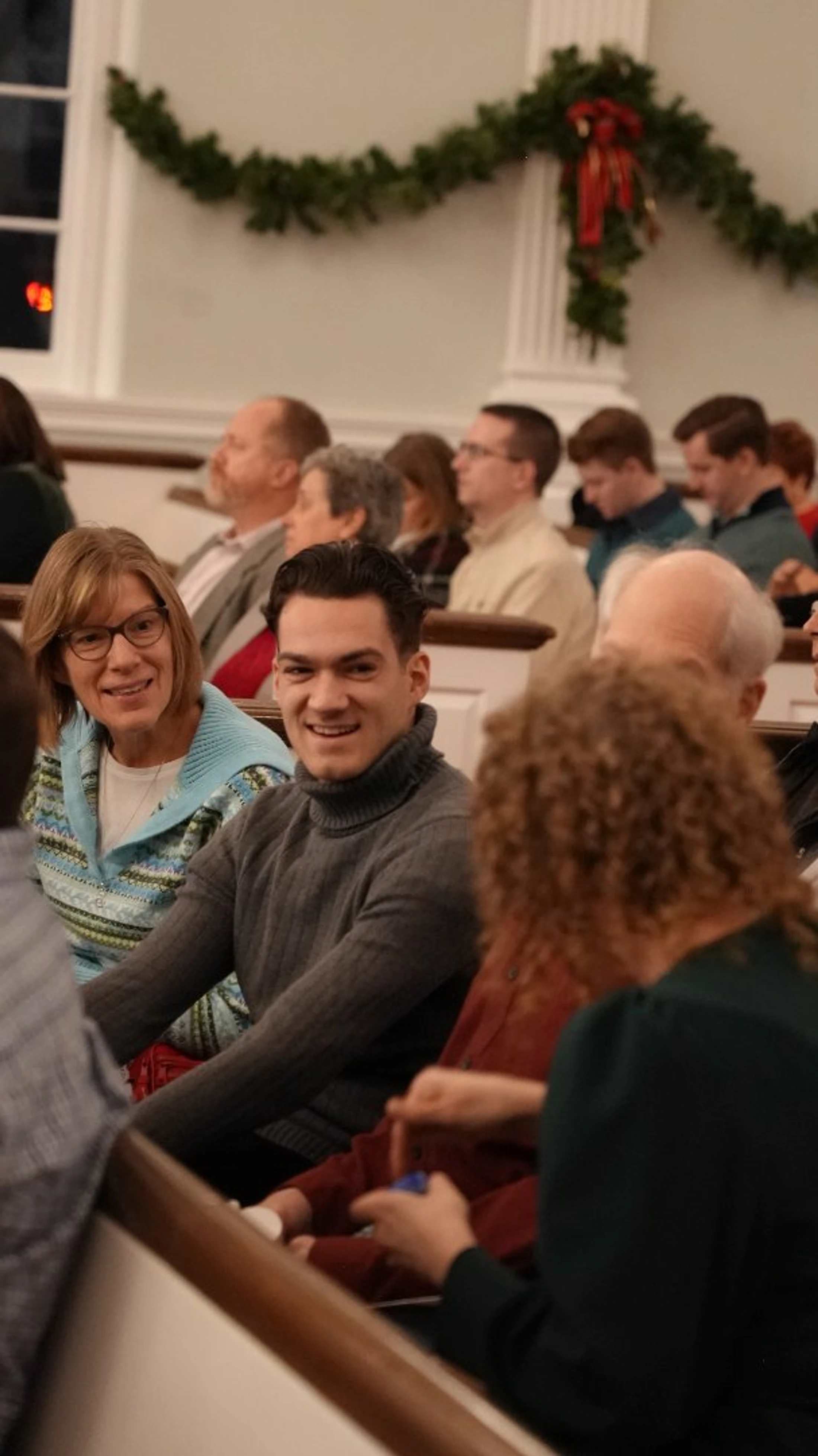 A young man in a grey turtleneck sweater smiles at the camera while sitting in a pew with other people during a holiday event in a church or hall.